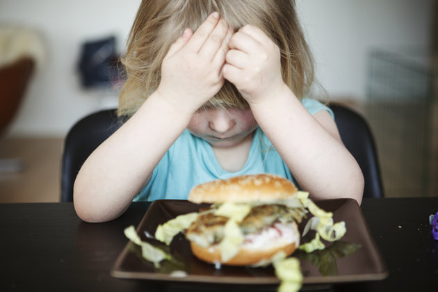 A three year old child/girl sitting by kitchen table w dinner/lunch hamburger in front of her. Head resting in hands, sour face, refusing to eat her food.