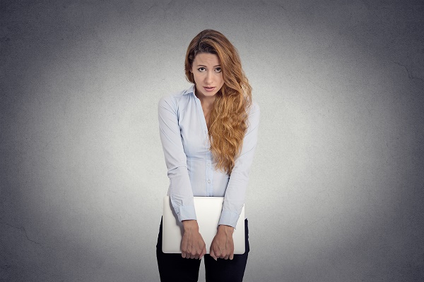 Lack of confidence. Insecure worried young woman holding laptop feels awkward isolated grey wall background. Human face expression emotion body language life perception