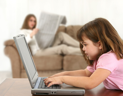 Five year old girl laying on table with laptop while mom (out of focus) reads newspaper on couch. Shot in studio.