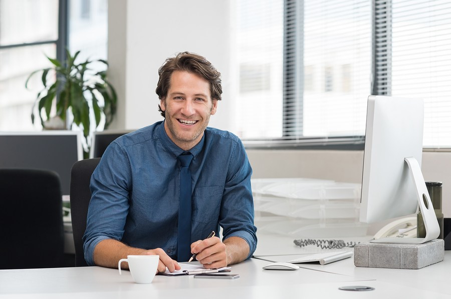 Cheerful young businessman working at desk in office. Successful business man sitting in office with a cup of coffee. Portrait of happy young businessman laughing and looking at camera.